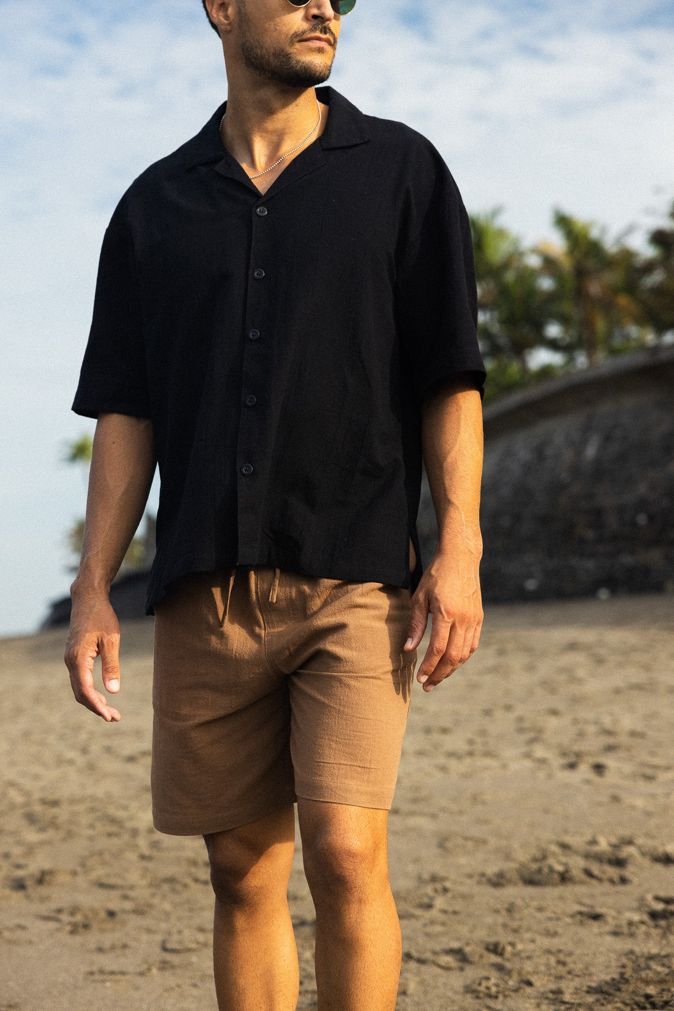 Man wearing a black shirt and beige shorts standing on a beach.